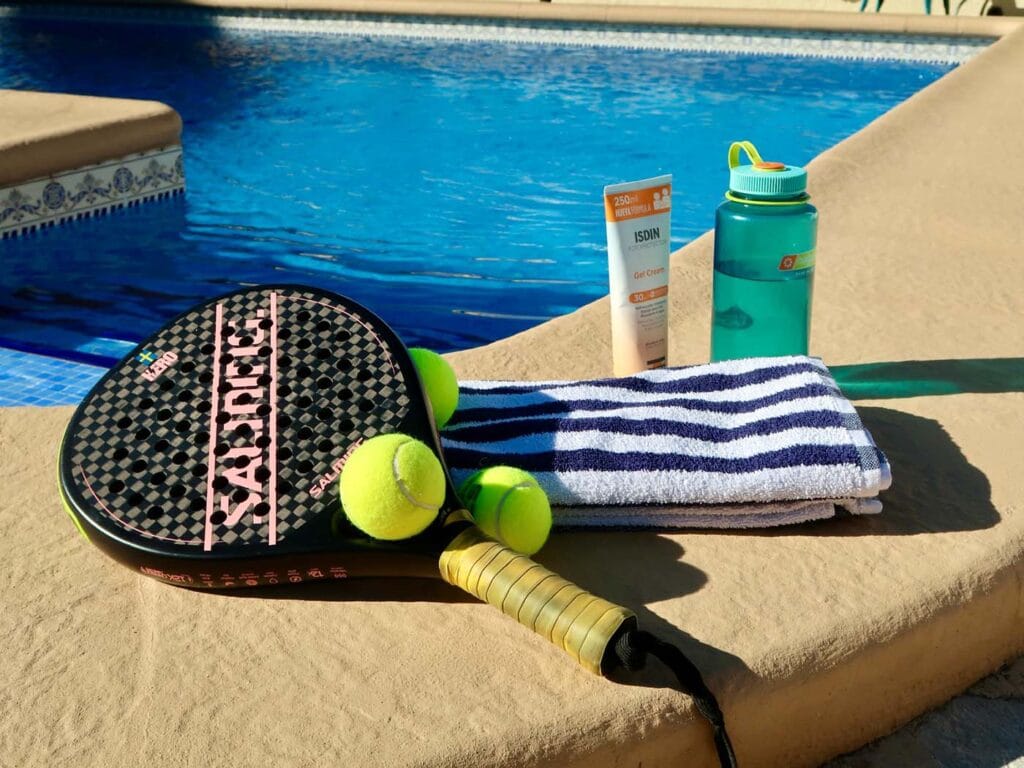 Padelracket, ballen, handdoek en zonnebrandcrème naast een zwembad; sfeerbeeld voor Casa Llibertat padelvakantie in Alicante.