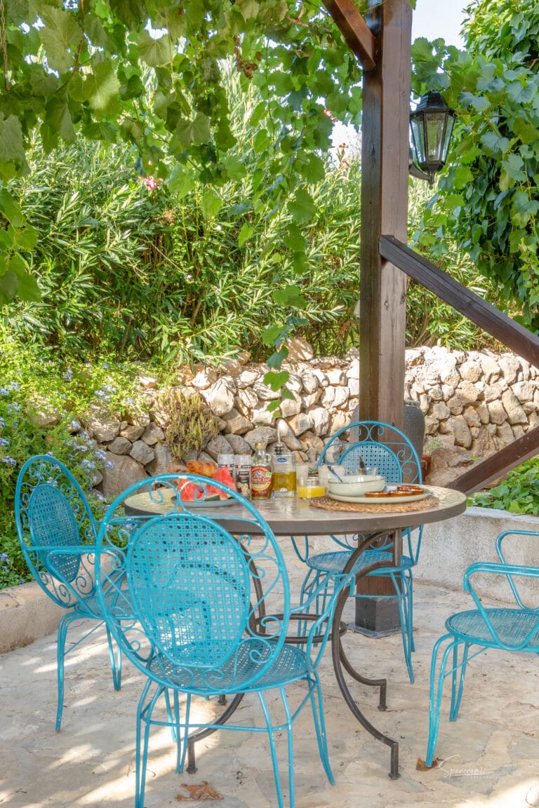 Kleine ronde tafel met turquoise stoelen op het buitenterras van Casa Libertat, omgeven door mediterrane beplanting.