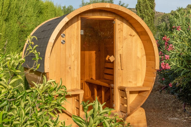 Una sauna de barril de madera en el jardín de Casa Llibertat en Llíber, rodeada de vegetación, ideal para relajarse durante unas vacaciones de pádel y bienestar.